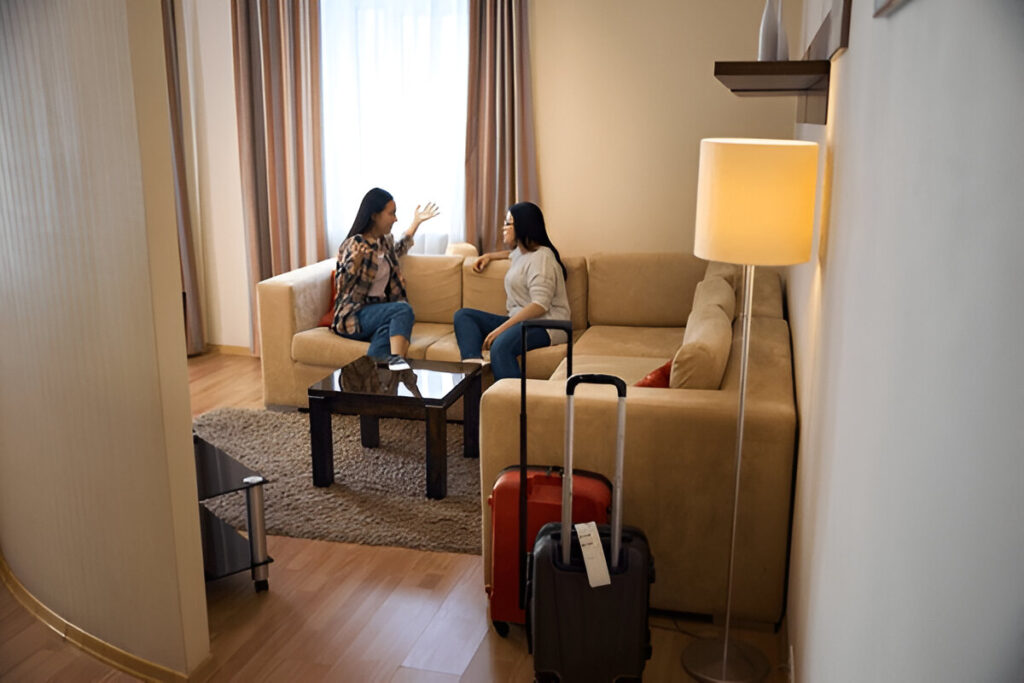 Two women talking in a modern living room with a beige sectional sofa and suitcases nearby