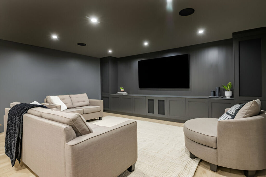 Modern home theater room with gray walls, built-in cabinets, a large flat-screen TV, and comfortable beige seating