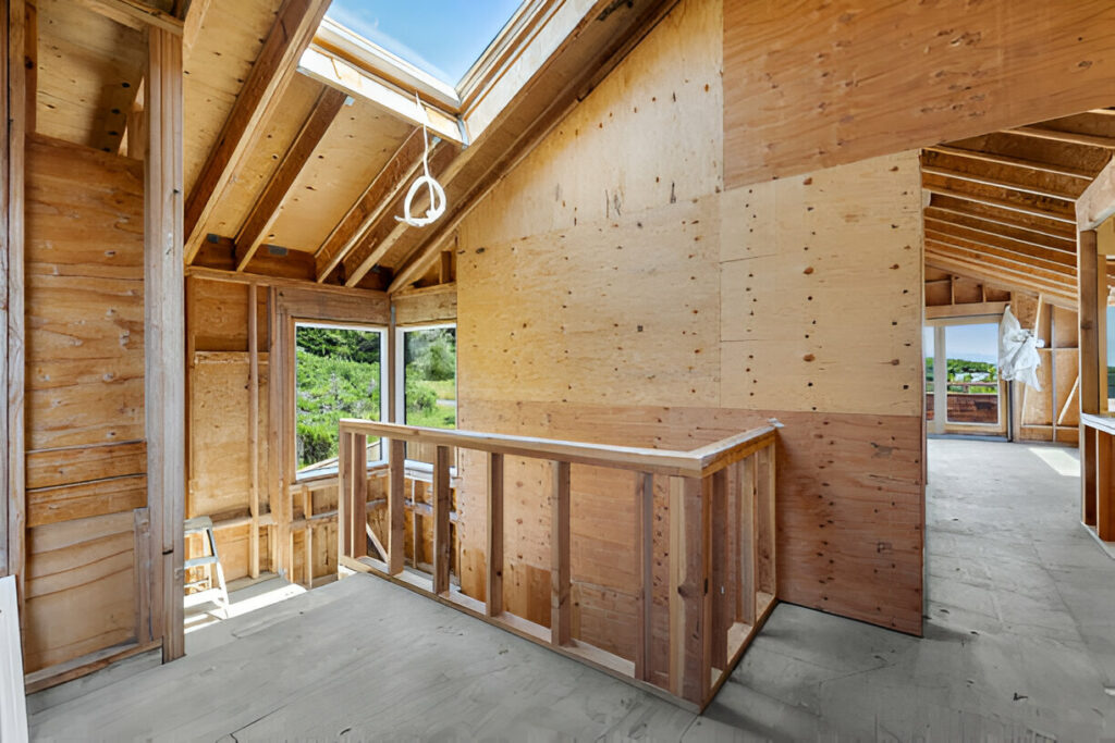 Interior of a home under construction with exposed wood framing, unfinished walls, and natural light coming through large windows and a skylight