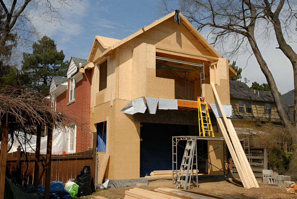 A two-story residential house under construction with exposed plywood sheathing, scaffolding, ladders, and construction materials in the front yard