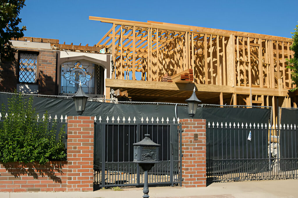 A residential property under renovation with a second-story wooden frame addition being built above the existing brick house, captured from the street view.