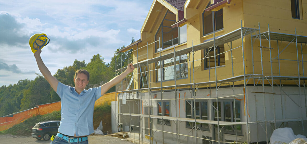 Happy construction worker celebrating in front of a partially built house with scaffolding and wooden exterior panels under a partly cloudy sky