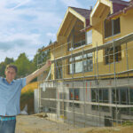 Happy construction worker celebrating in front of a partially built house with scaffolding and wooden exterior panels under a partly cloudy sky