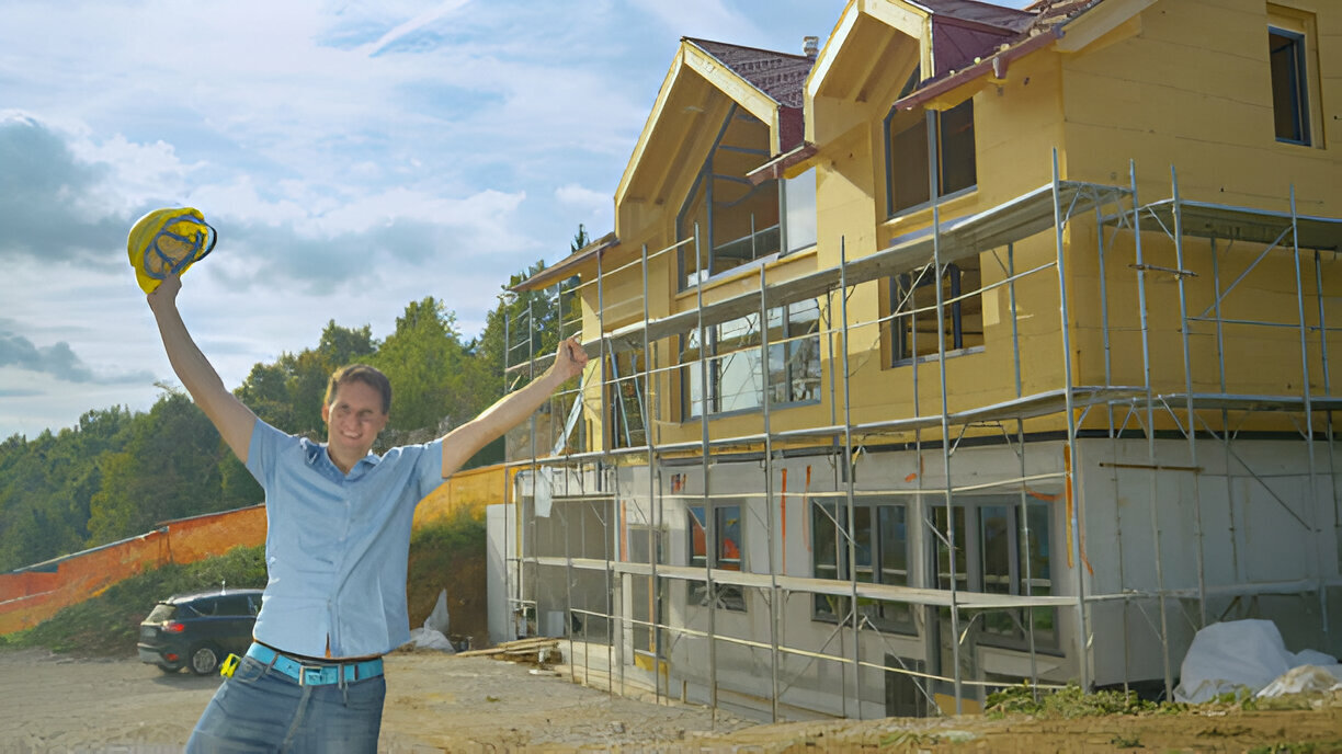 Happy construction worker celebrating in front of a partially built house with scaffolding and wooden exterior panels under a partly cloudy sky