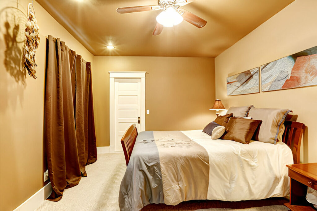 Warm and inviting bedroom with earth-toned walls, a wooden bed frame, plush bedding, and decorative pillows. The room features a ceiling fan with a light fixture, rich brown curtains, and modern artwork on the wall.

