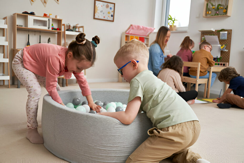 Children playing in a bright, modern playroom filled with educational toys, creative art stations, and a soft ball pit. The room features light wood furniture, cheerful decorations, and plenty of space for creative activities