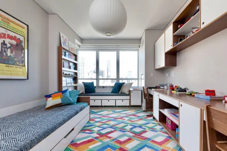 A bright and colorful multipurpose kids' room featuring a bed, desk, bookshelves, and a cozy window seat with storage underneath. The room has modern white and wood furniture, a colorful geometric rug, and large windows offering a city view