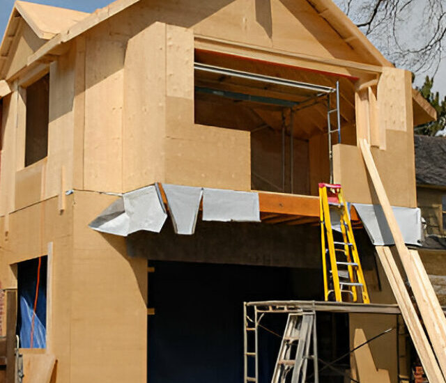 A two-story residential house under construction with exposed plywood sheathing, scaffolding, ladders, and construction materials in the front yard