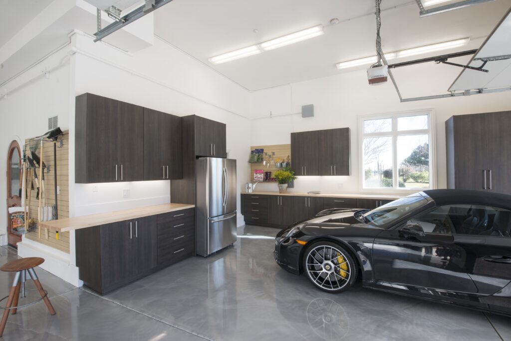 High-end residential garage featuring custom dark wood cabinetry, a sleek polished concrete floor, stainless steel appliances, and a luxury sports car.

