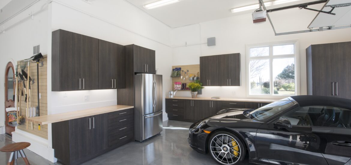High-end residential garage featuring custom dark wood cabinetry, a sleek polished concrete floor, stainless steel appliances, and a luxury sports car.