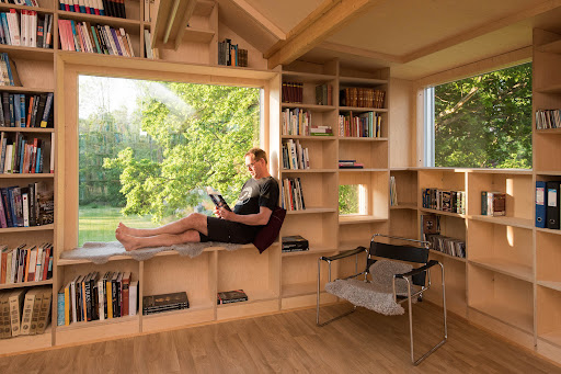 A cozy home library with custom-built wooden bookshelves, a large picture window with a reading bench, and a man relaxing with a book while enjoying views of lush greenery outside.

