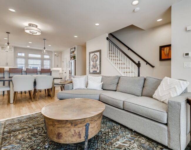 Open-concept living space with light wood floors, gray sectional sofa, rustic round coffee table, dining area, and white kitchen in the background.