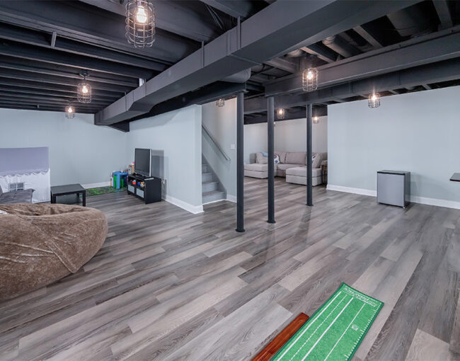 Open-concept basement with exposed black ceiling beams, vinyl plank flooring, lounge area, kids’ play zone, and putting green.