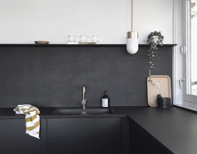 Minimalist kitchen with matte black cabinets and backsplash, gold faucet, white pendant light, and modern decor elements.