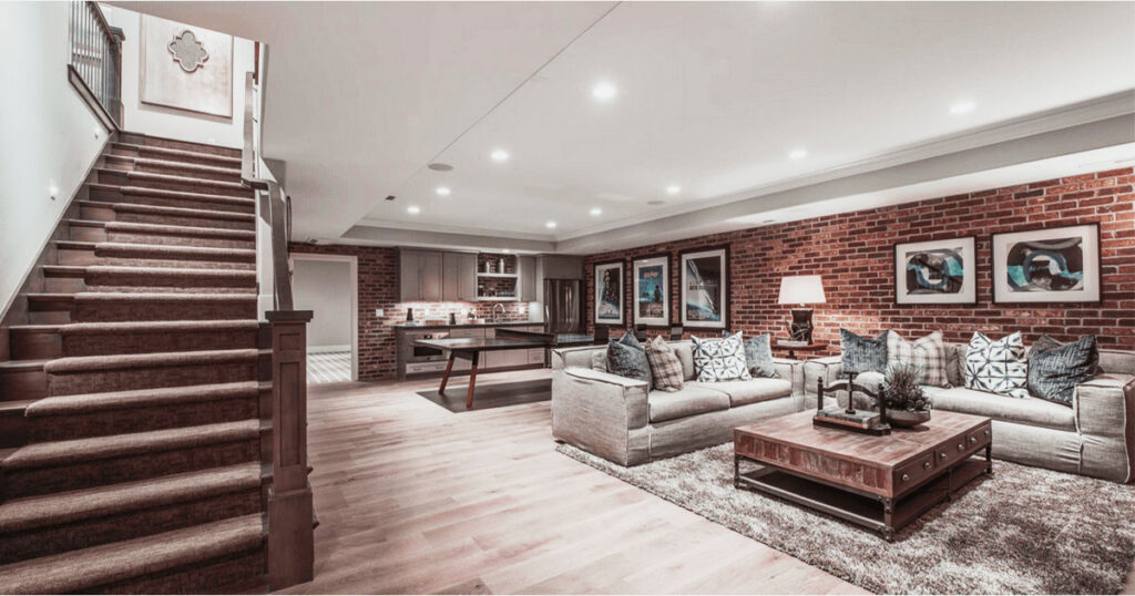 Finished basement with exposed red brick walls, built-in kitchenette, plush seating area, wooden staircase, and cozy lighting.