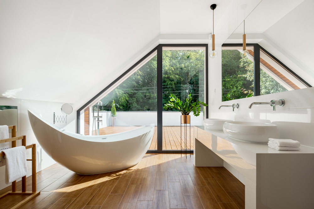 The image shows a spacious bathroom with a freestanding bathtub, double vanity sinks, and large windows overlooking a lush outdoor landscape. The design features a harmonious blend of natural wood tones and clean white surfaces