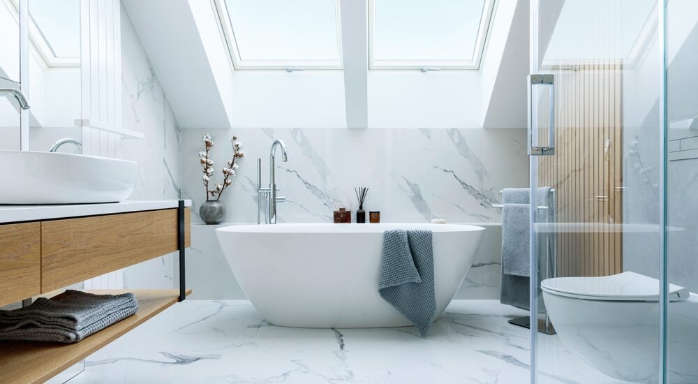 The image shows a stunning bathroom with a freestanding bathtub, a double vanity, and a glass-enclosed shower. The space is characterized by a sleek, modern design featuring extensive use of white marble and natural wood accents.