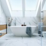The image shows a stunning bathroom with a freestanding bathtub, a double vanity, and a glass-enclosed shower. The space is characterized by a sleek, modern design featuring extensive use of white marble and natural wood accents.