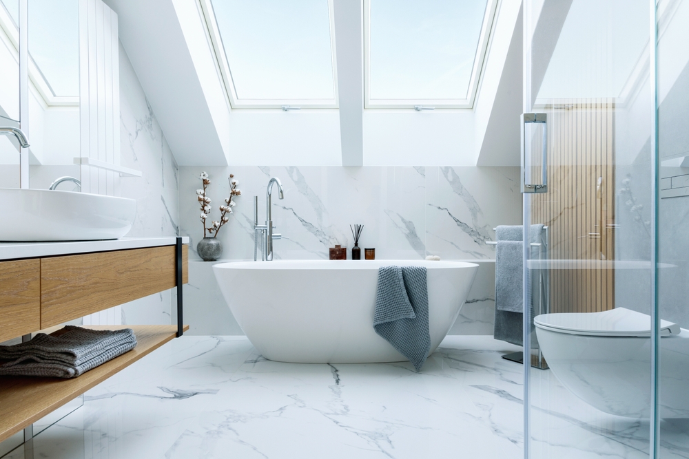 The image shows a stunning bathroom with a freestanding bathtub, a double vanity, and a glass-enclosed shower. The space is characterized by a sleek, modern design featuring extensive use of white marble and natural wood accents.