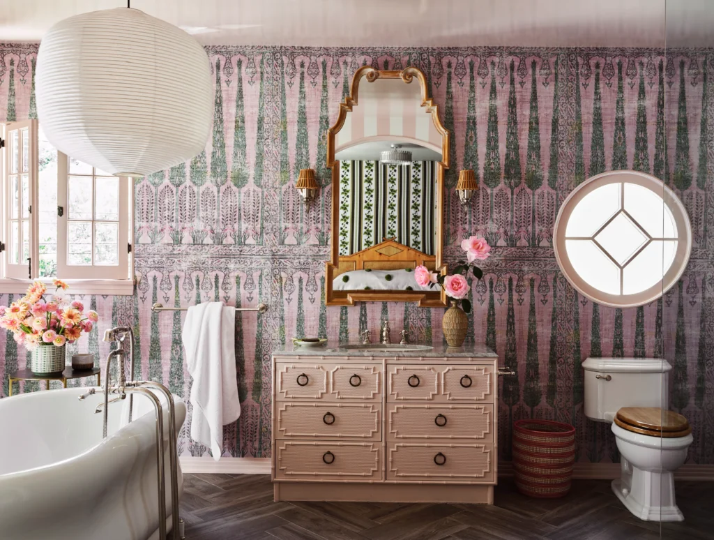 Luxurious bathroom with patterned pink wallpaper, ornate pink vanity, sculptural mirror, and circular window creating a romantic vintage vibe.