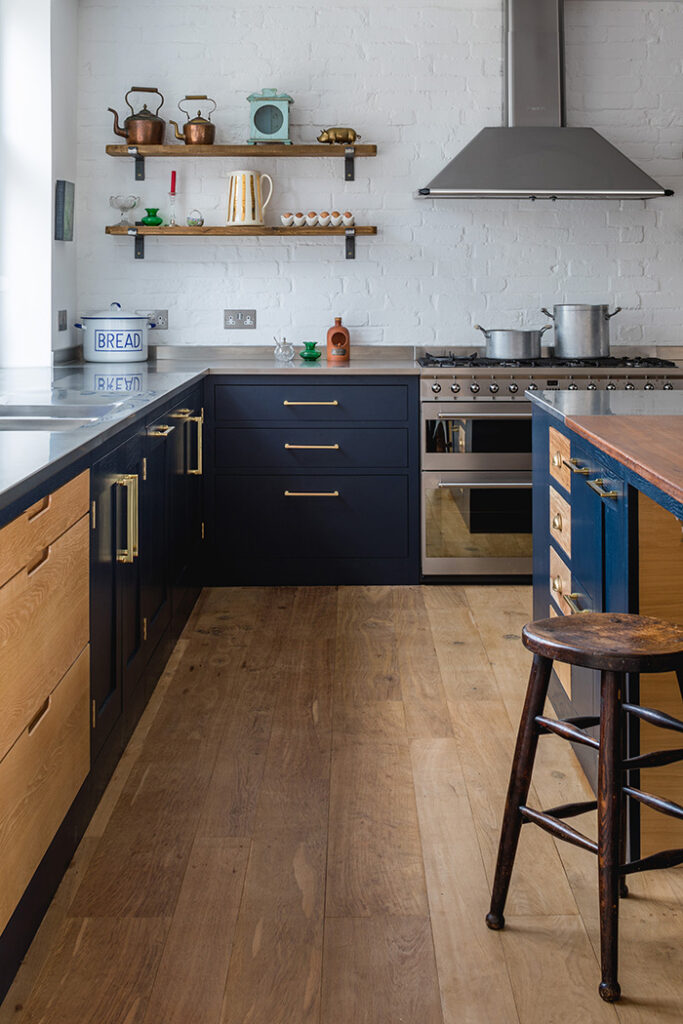 Creative Kitchen Floor Ideas 10 Rustic kitchen with Victorian beam oak wood flooring, navy and natural wood cabinetry, brass hardware, and exposed white brick walls.