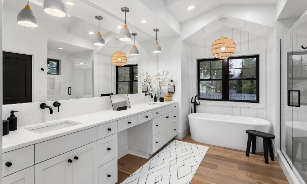 Custom Cabinetry Trends for Bathrooms to Watch in 2025 7 Spacious bathroom featuring a freestanding soaking tub, long white vanity with black fixtures, pendant lights, and wood flooring