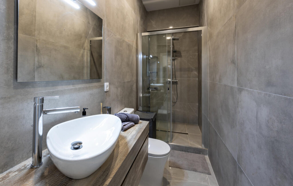 Modern small bathroom with gray large-format tiles, floating wooden vanity, white vessel sink, wall mirror, and a glass-enclosed shower.
