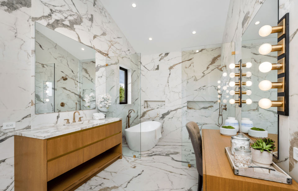 Custom Cabinetry Trends for Bathrooms to Watch in 2025 16 Elegant bathroom with full marble walls and flooring, wood vanity, freestanding tub, and lighted makeup station