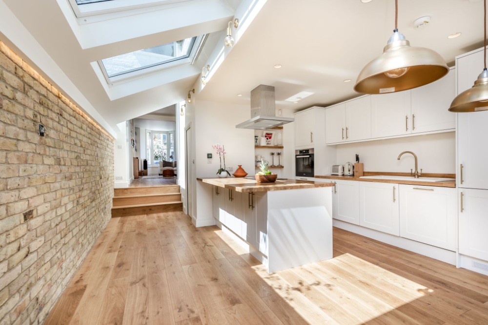 Top Kitchen Flooring Trends for 2025 8 Modern white kitchen with wood countertops, island, oak flooring, exposed brick wall, and skylights bringing in natural light.
