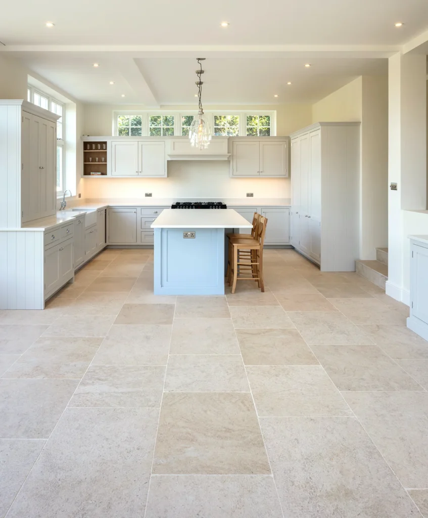 The Ultimate Guide to Kitchen and Floor Tiles 10 Spacious kitchen with light beige limestone tile flooring, pastel gray cabinets, central island with wooden stools, and modern lighting