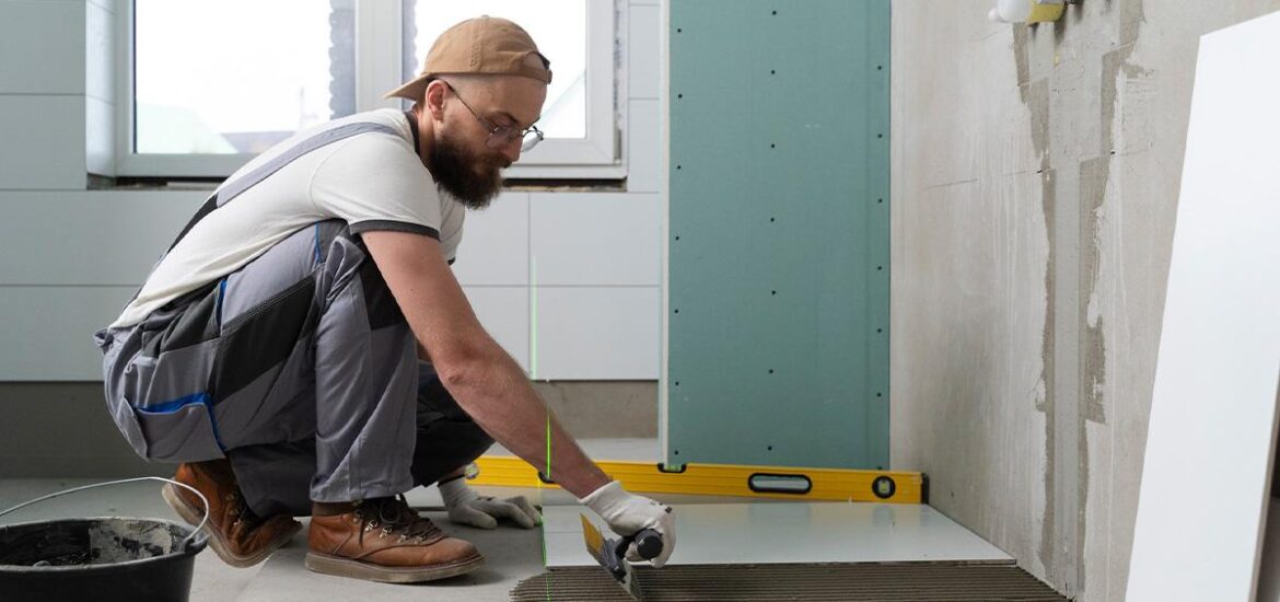 A construction worker in overalls and gloves installing a large floor tile in a partially renovated bathroom, using adhesive and a notched trowel.