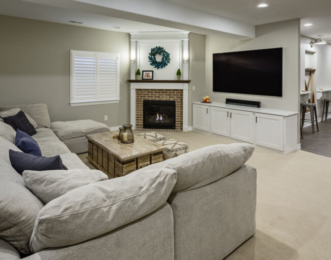 Finished basement featuring a large sectional sofa, rustic wood coffee table, brick fireplace, and wall-mounted TV