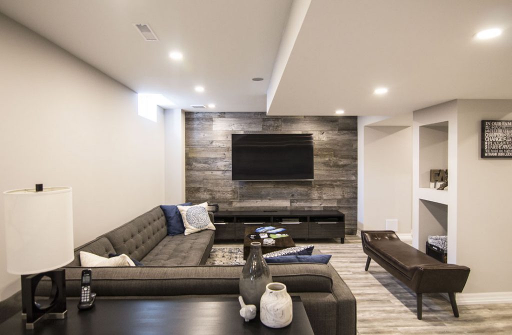Cozy basement living area with gray sectional, wood accent wall, mounted TV, and recessed lighting