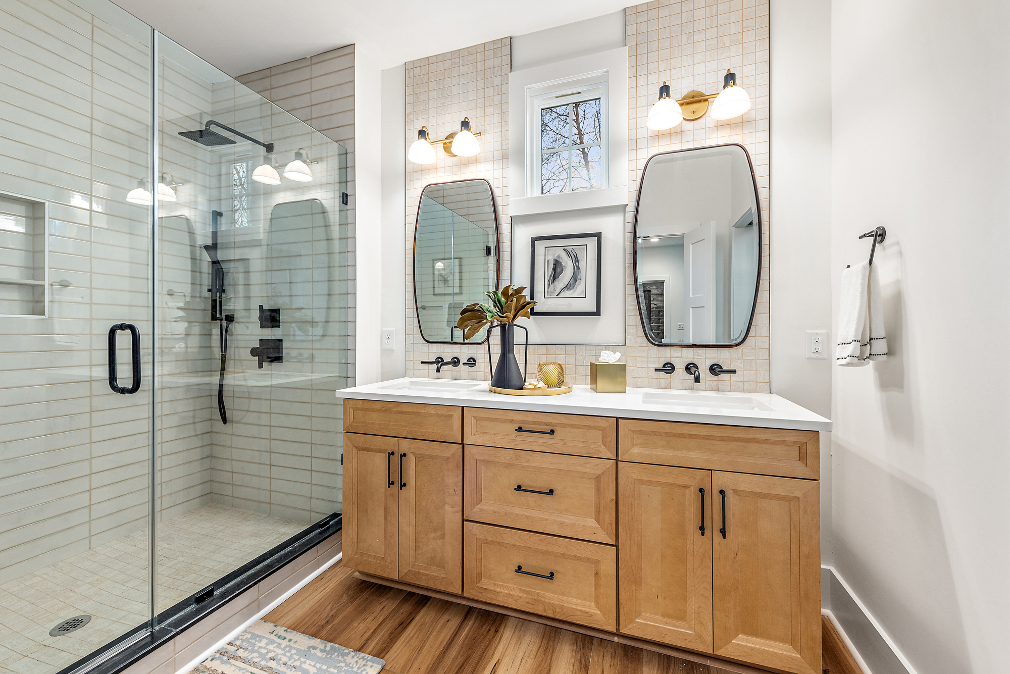Modern bathroom with a glass-enclosed shower, dual vanity with wooden cabinets, and two framed mirrors under wall-mounted lights.