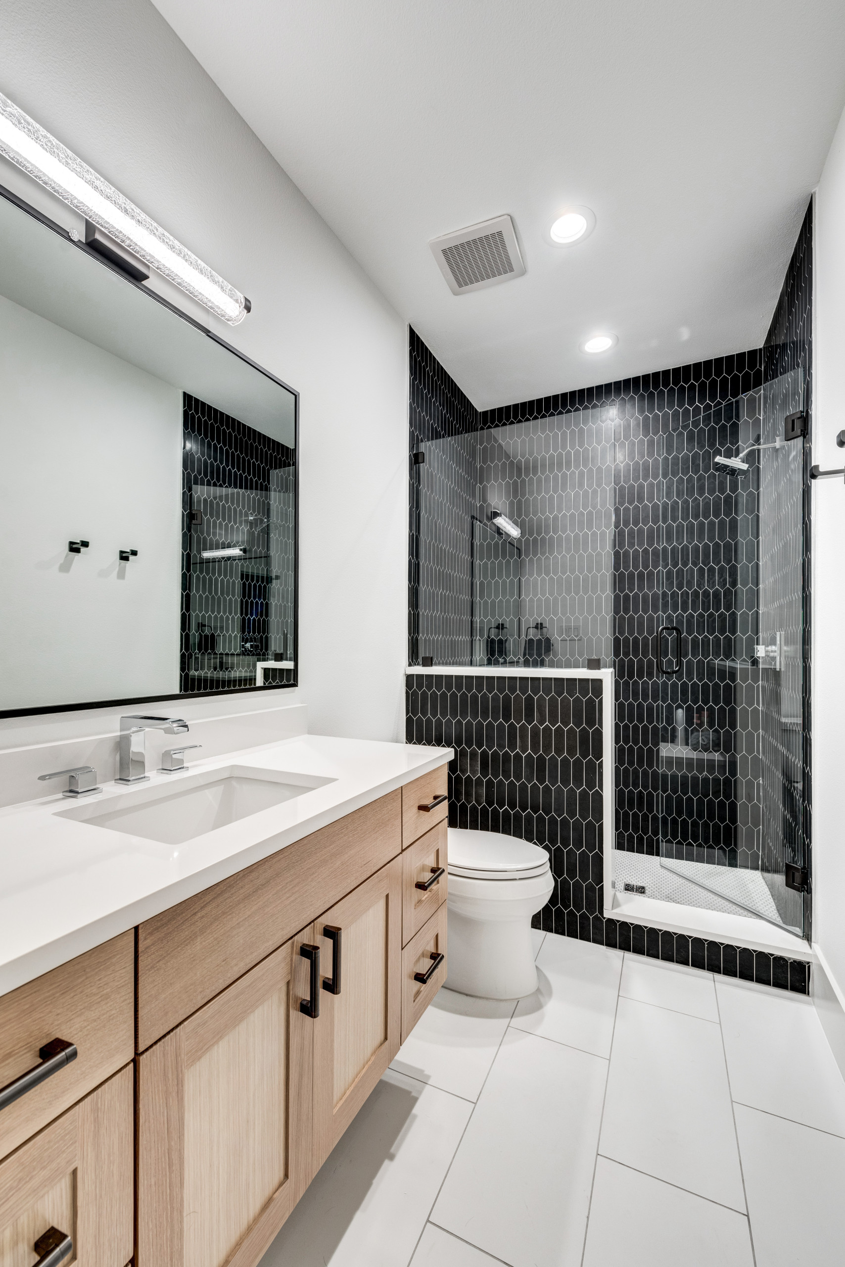 Modern bathroom with light wood vanity, black hexagonal wall tiles, and glass shower enclosure.
