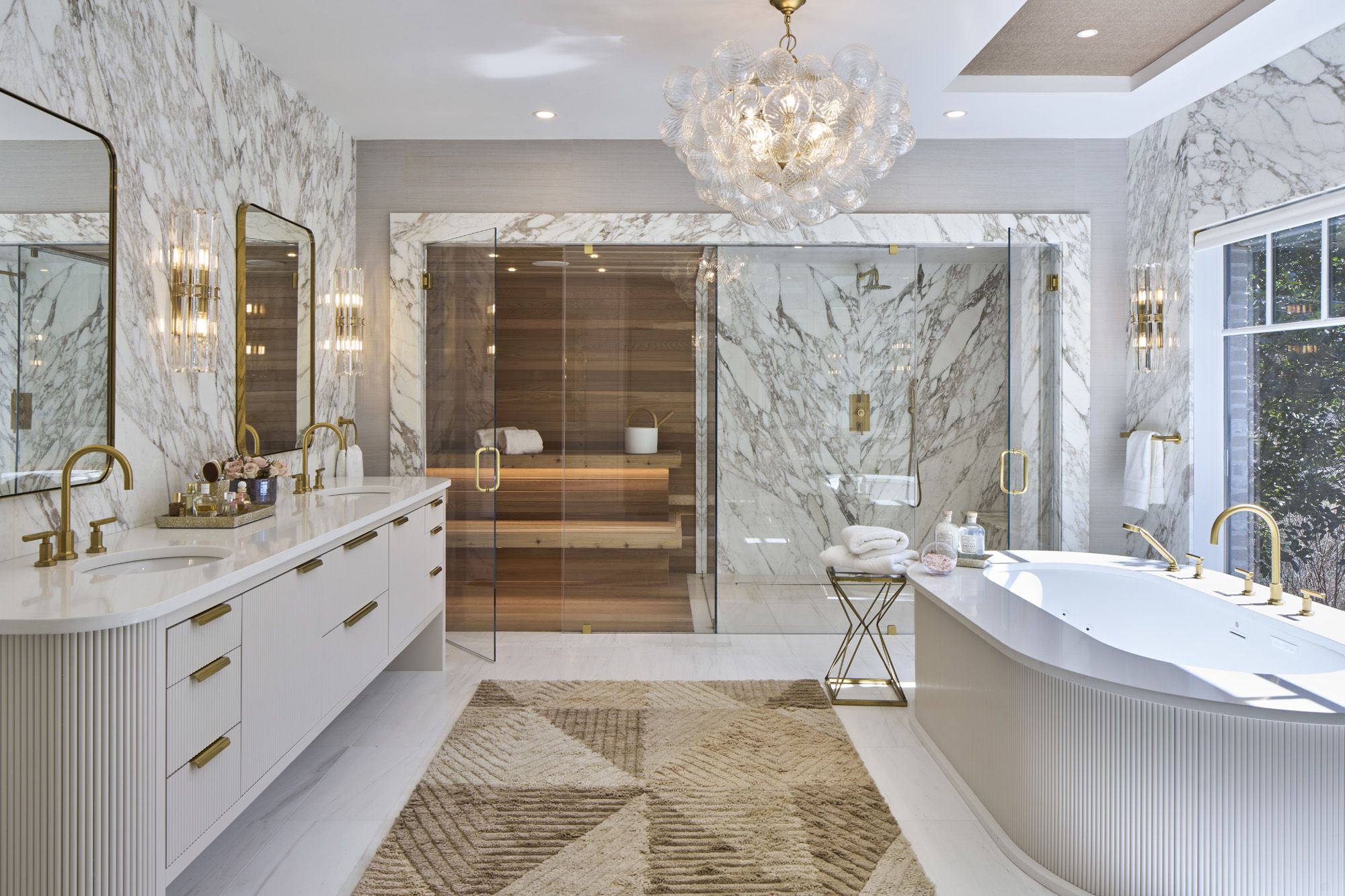 Luxurious marble bathroom with gold fixtures, freestanding tub, double vanity, glass-enclosed shower, and sauna.