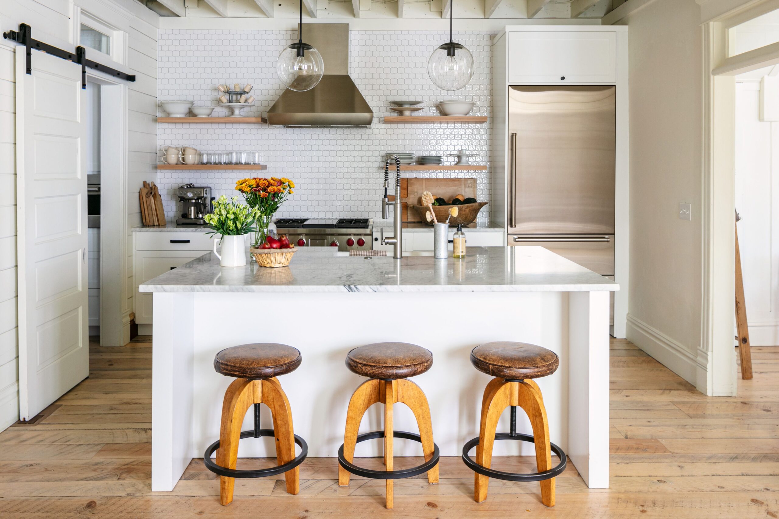 Bright farmhouse kitchen with a marble island, wooden stools, open shelving, and stainless steel appliances.