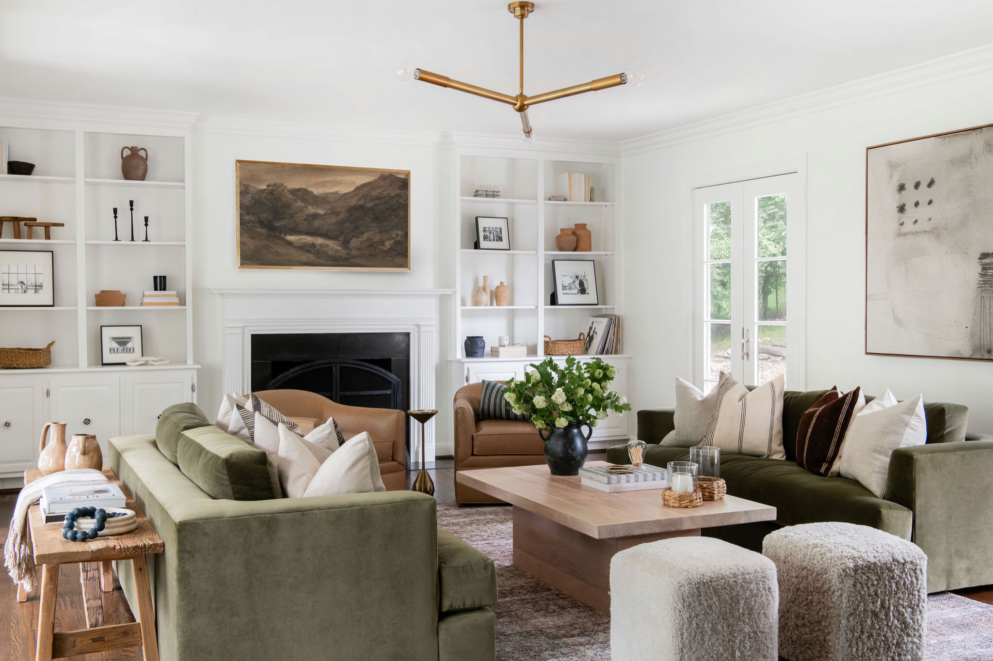 Bright and elegant living room with green velvet sofas, neutral-toned armchairs, a light wood coffee table, and built-in white shelves surrounding a fireplace.