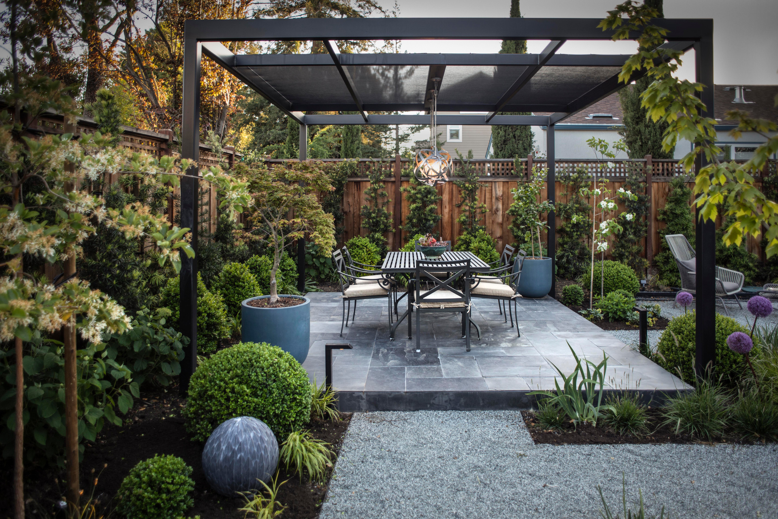 A contemporary backyard dining area with a black pergola, a sleek metal dining set, and a modern pendant light. Surrounded by lush greenery, manicured shrubs, and decorative planters, the space is set on a stone patio with gravel pathways.