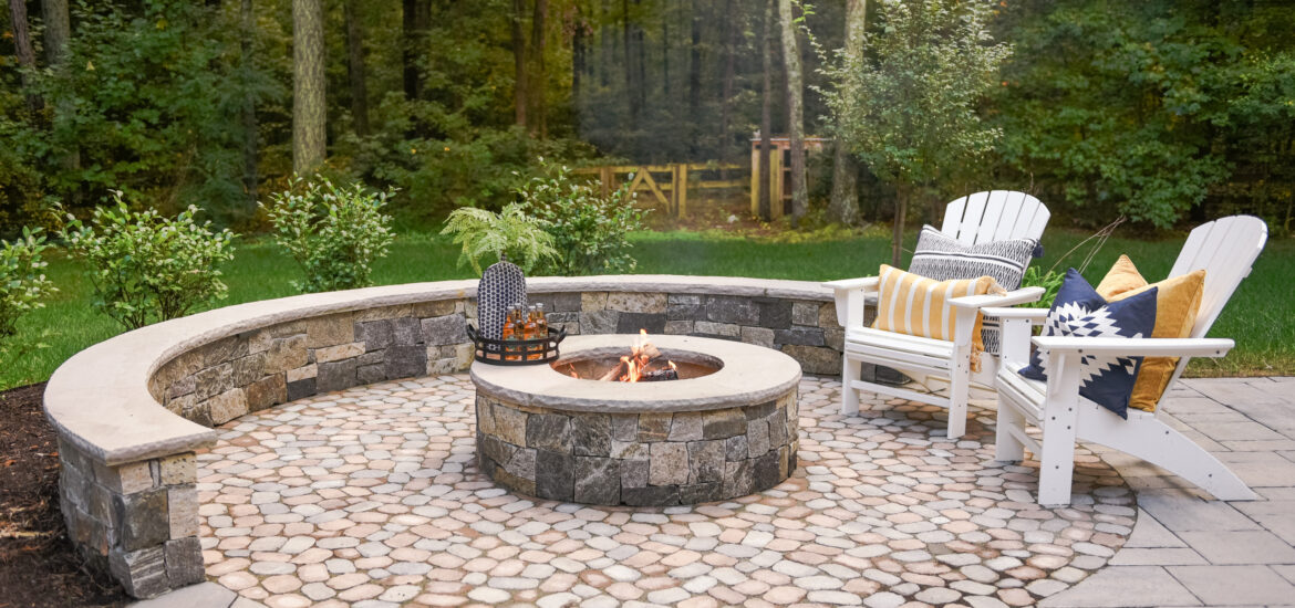 A rustic outdoor fire pit area featuring a circular stone patio with a built-in stone bench surrounding a central fire pit. Two white Adirondack chairs with colorful cushions and pillows add comfort, while the backdrop of dense forest creates a serene atmosphere.