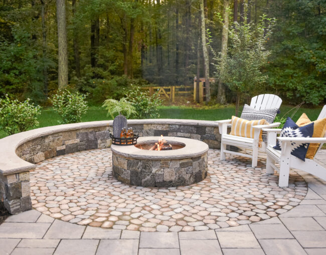 A rustic outdoor fire pit area featuring a circular stone patio with a built-in stone bench surrounding a central fire pit. Two white Adirondack chairs with colorful cushions and pillows add comfort, while the backdrop of dense forest creates a serene atmosphere.