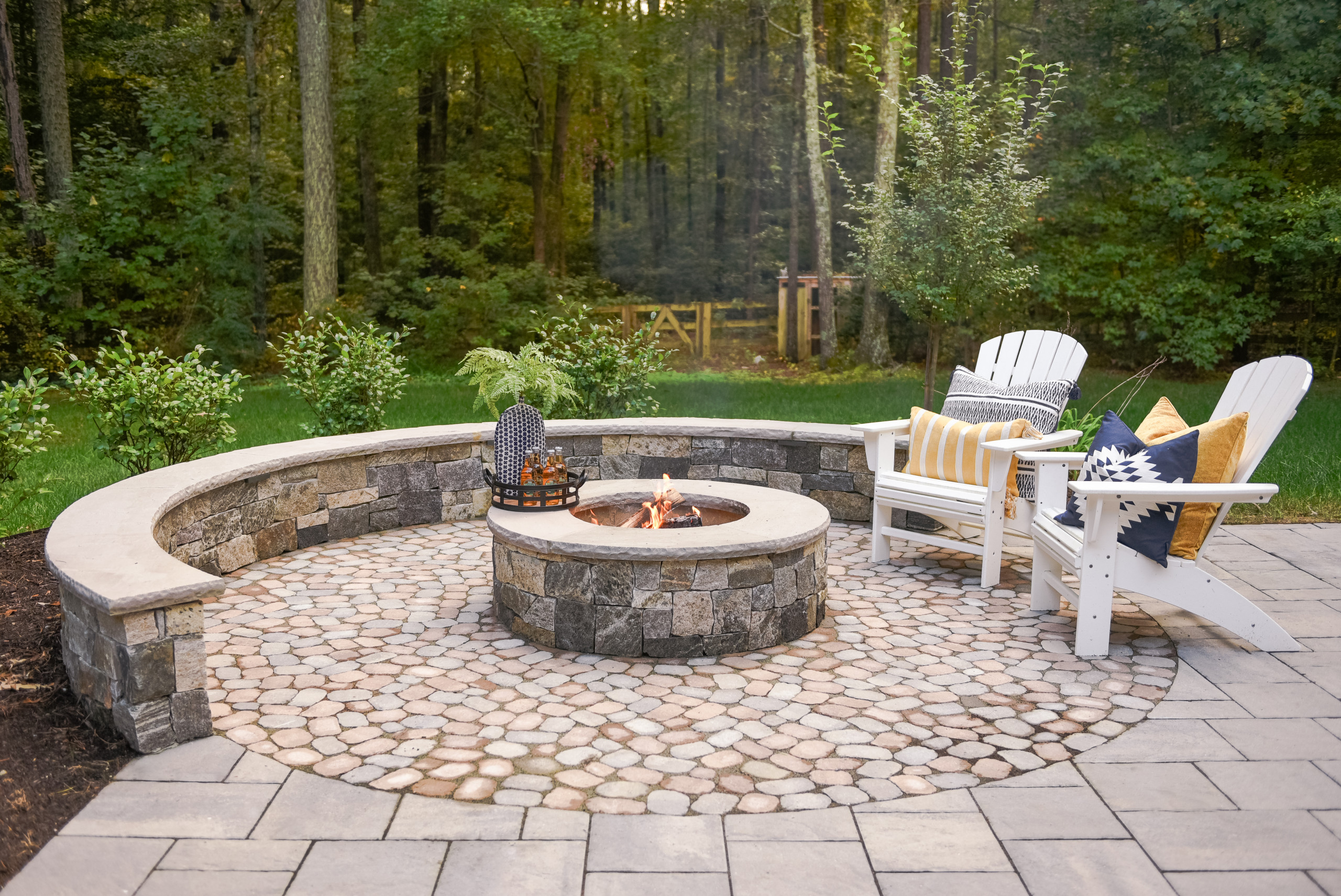 A rustic outdoor fire pit area featuring a circular stone patio with a built-in stone bench surrounding a central fire pit. Two white Adirondack chairs with colorful cushions and pillows add comfort, while the backdrop of dense forest creates a serene atmosphere.