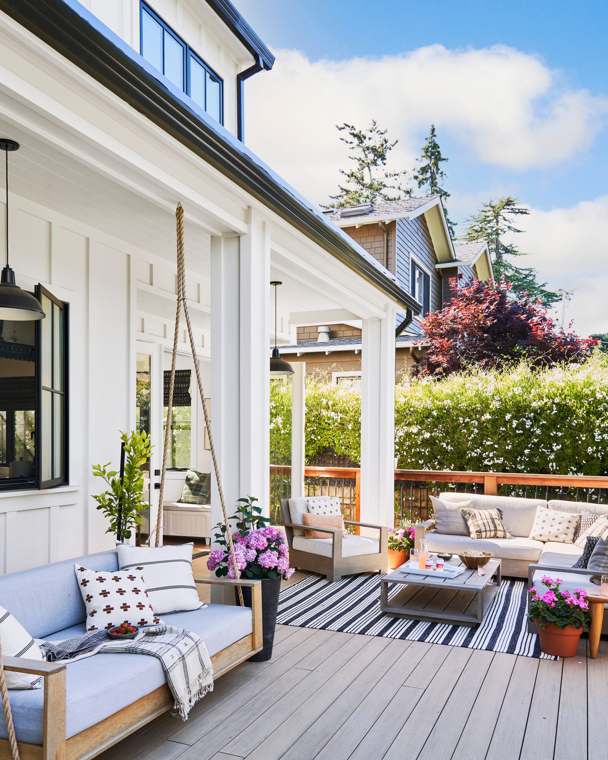 Bright outdoor patio with comfortable seating, decorative pillows, striped rug, potted flowers, and a cozy atmosphere under a covered porch.