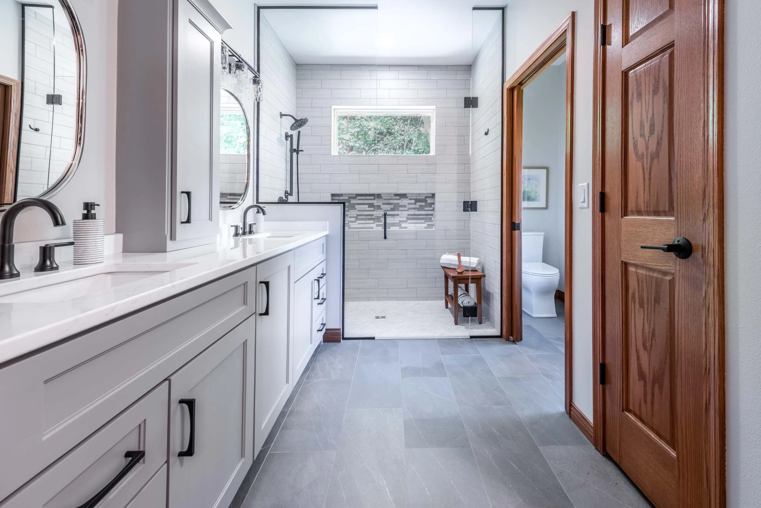 Modern bathroom with white vanity, black fixtures, large mirrors, and a walk-in shower with gray subway tiles and accent mosaic strip.