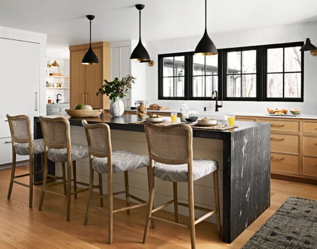 Modern kitchen with black marble waterfall island, natural wood accents, and woven bar stools under black pendant lights.