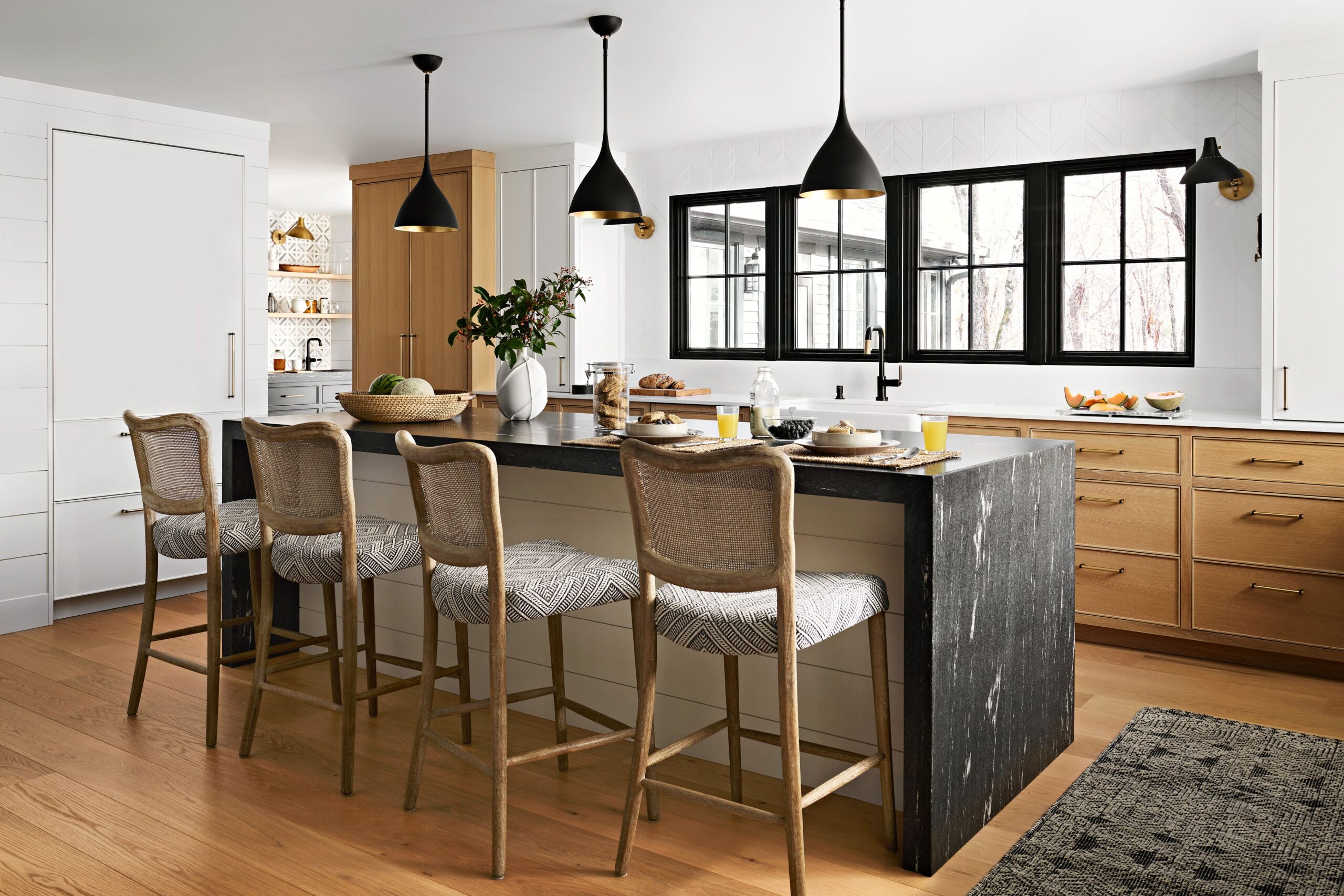 Modern kitchen with black marble waterfall island, natural wood accents, and woven bar stools under black pendant lights.