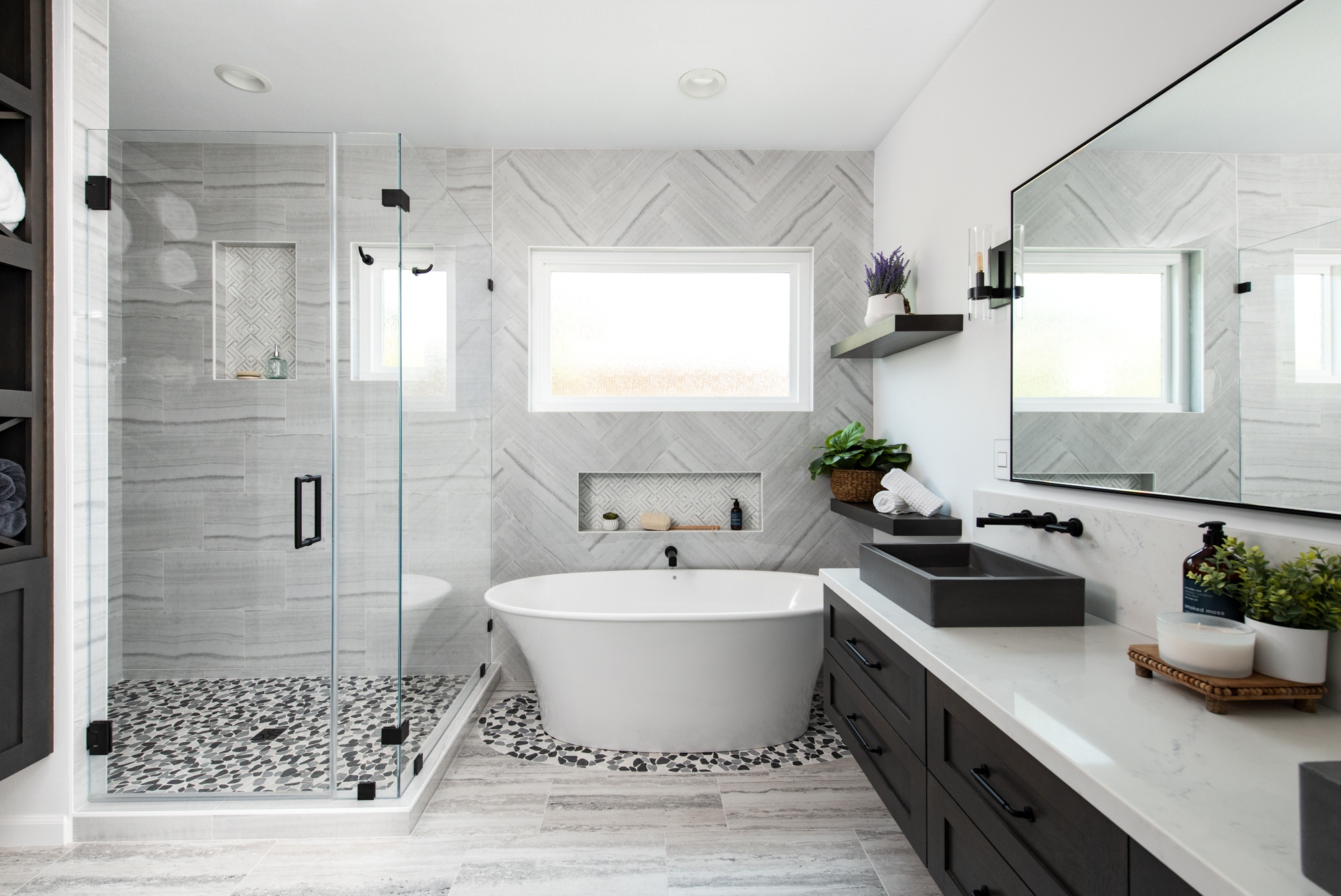 Modern master bathroom with freestanding bathtub, glass-enclosed shower, herringbone-pattern wall tiles, and dark wood vanity with vessel sinks.
