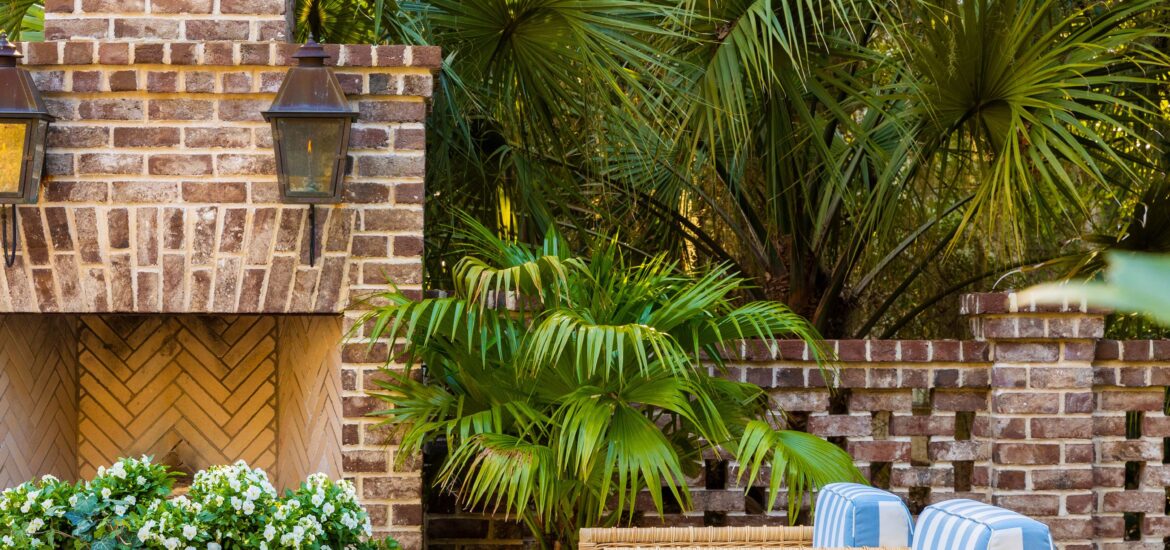 Outdoor patio with brick fireplace, wicker chairs with striped cushions, tropical greenery, and a white coffee table with fresh flowers.