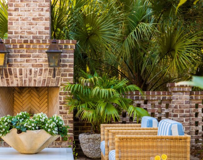 Outdoor patio with brick fireplace, wicker chairs with striped cushions, tropical greenery, and a white coffee table with fresh flowers.