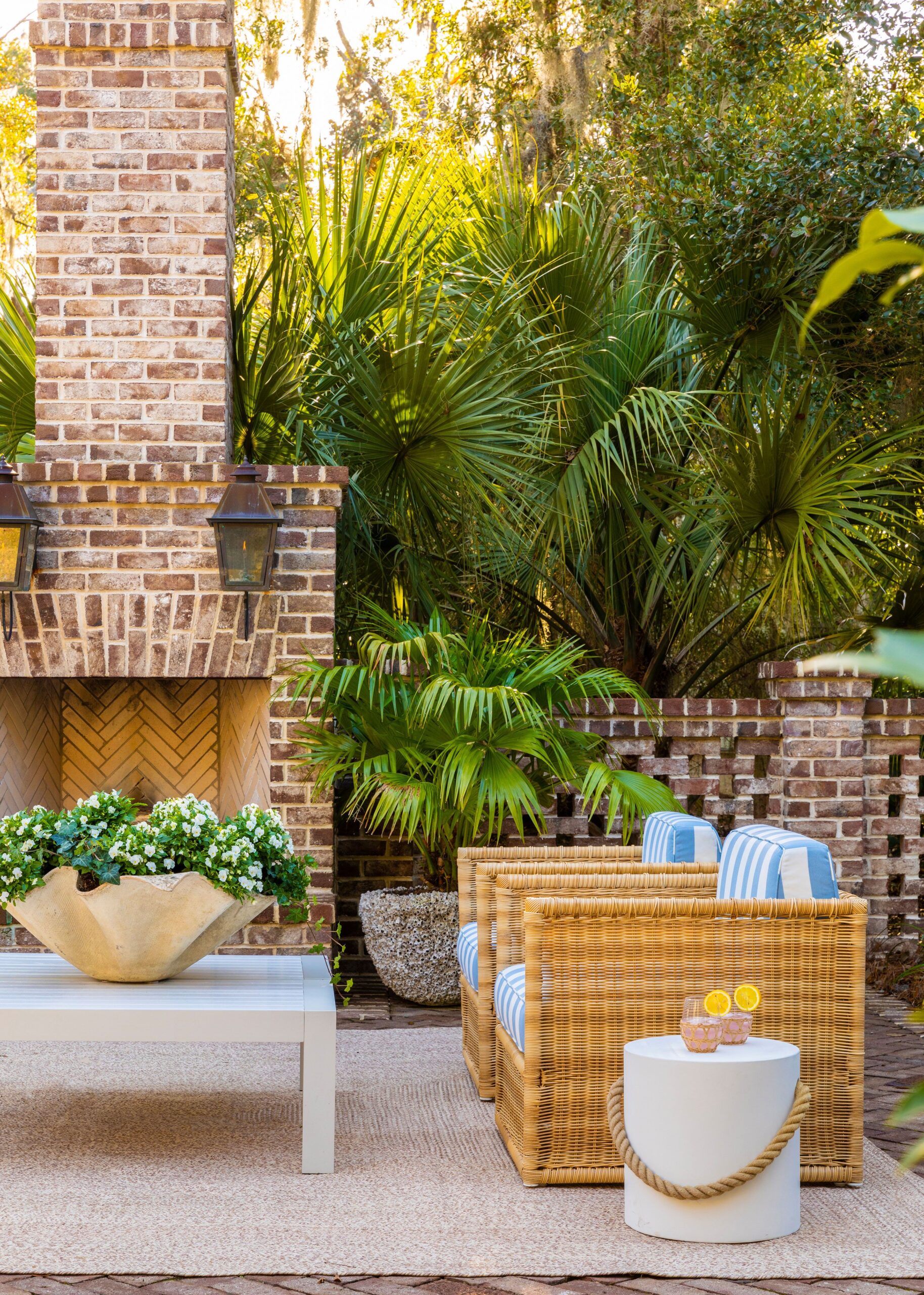 Outdoor patio with brick fireplace, wicker chairs with striped cushions, tropical greenery, and a white coffee table with fresh flowers.